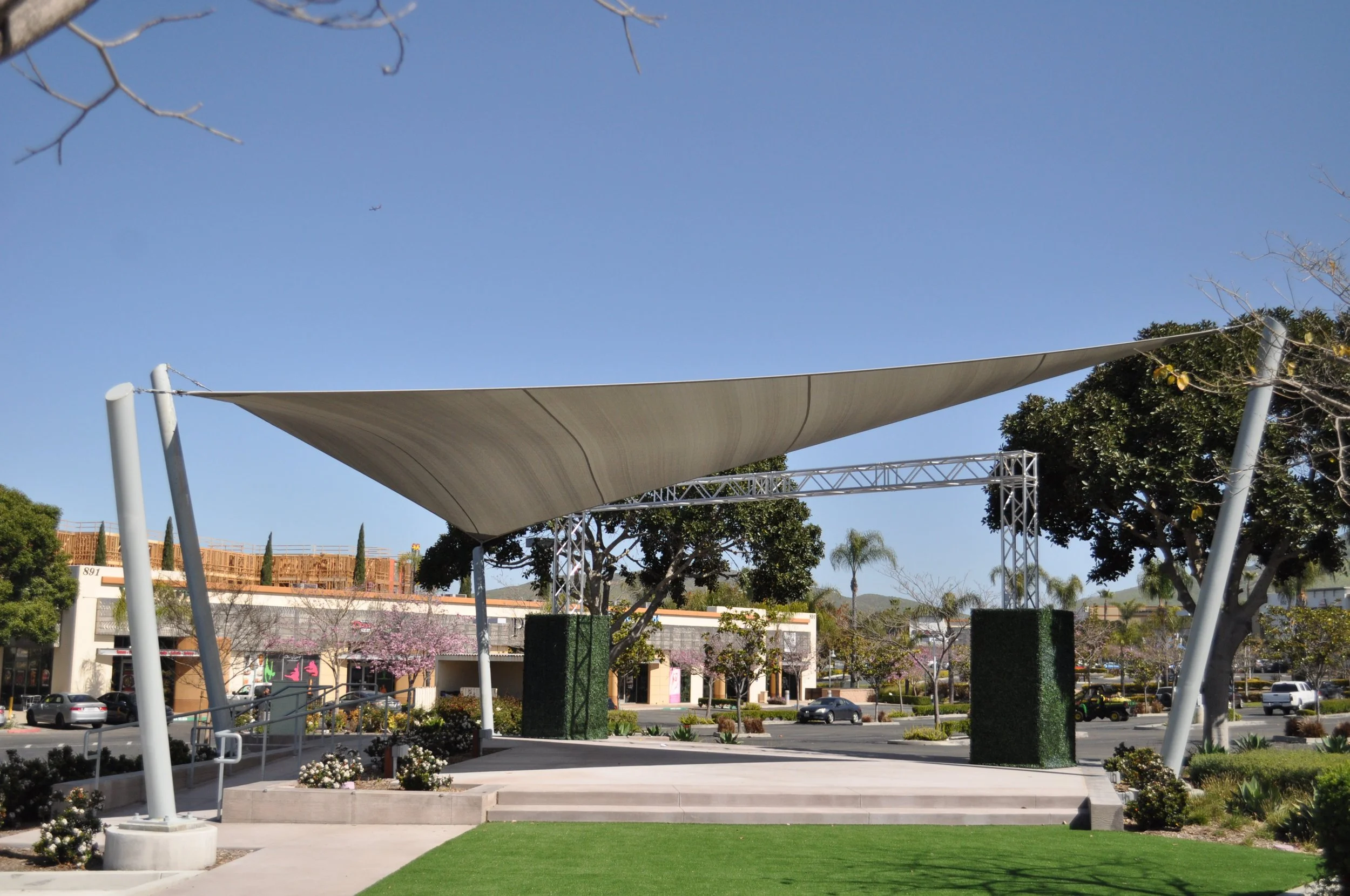 A modern outdoor stage with a fabric canopy, supported by angled poles, in a landscaped area with trees, flowers, and a parking lot in the background.