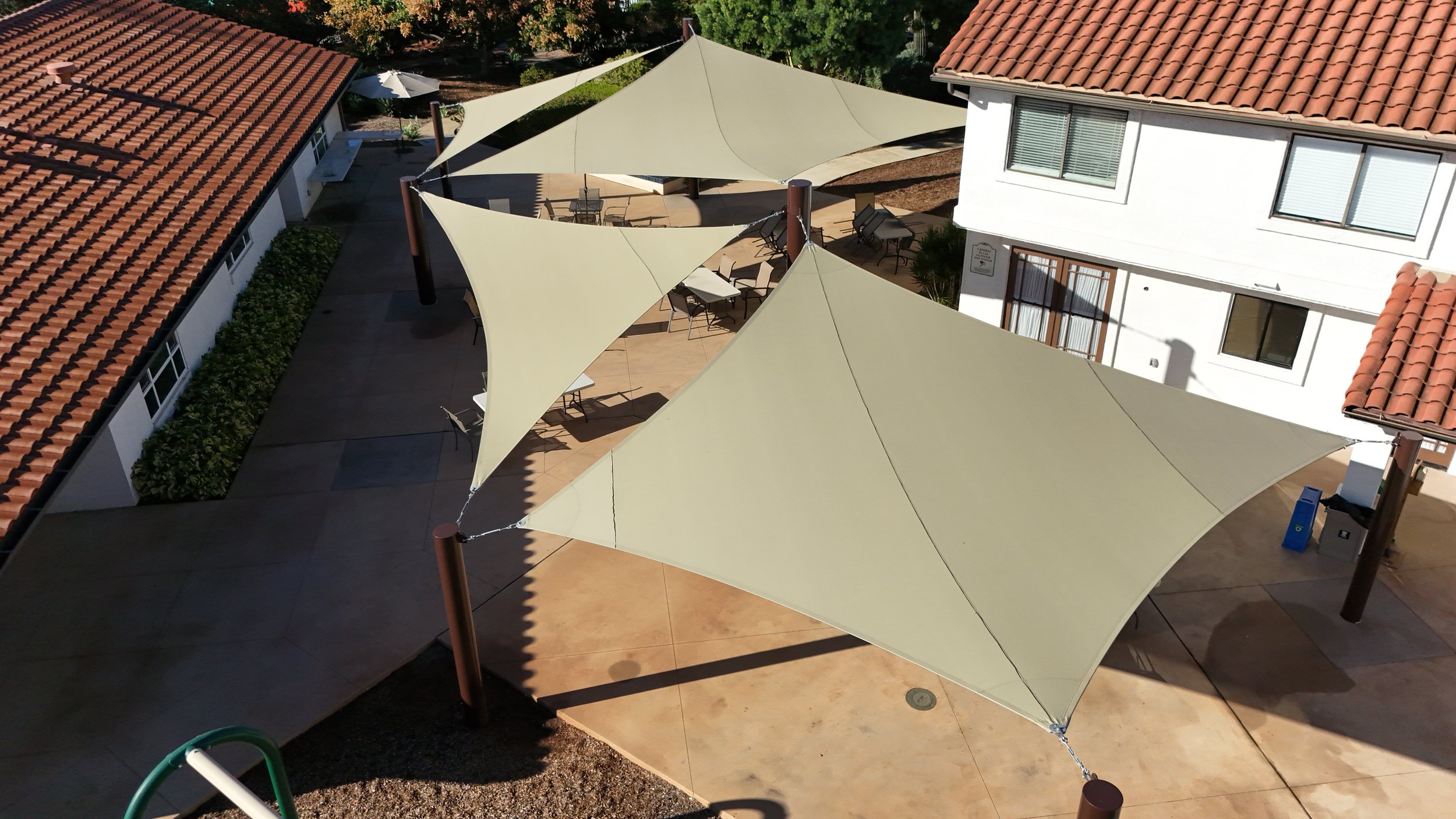 Aerial view of an outdoor patio area with beige tensile shade sails supported by wooden poles, surrounding white buildings with red-tile roofs, tables and chairs underneath the shade sails.