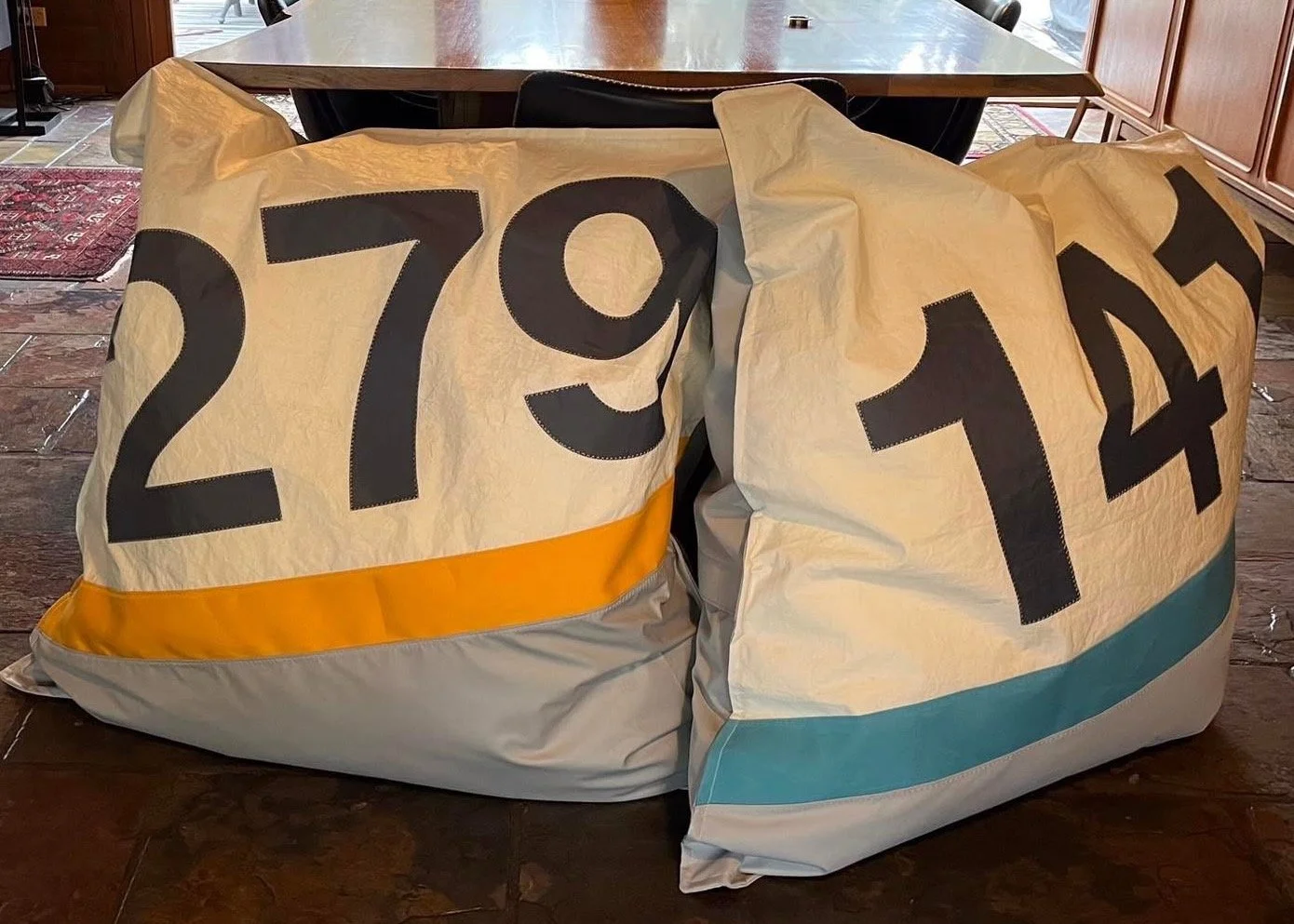 Large fabric bags with bold black numbers, possibly used for storage or decoration, on a wooden indoor floor with a table and chairs in the background.