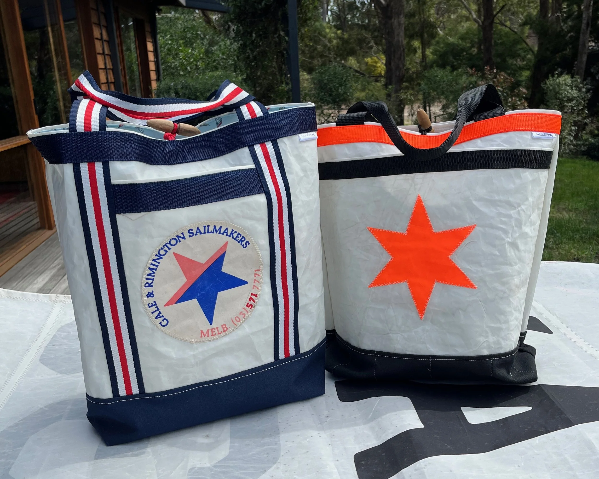 Two tote bags with sailboat-themed logos on a white table outside. One bag has a blue, red, and white color scheme with a round logo featuring a star and the text 'Clyde & Kimmington Sailmakers MELB.' The other bag has a red star logo and black and orange trim.