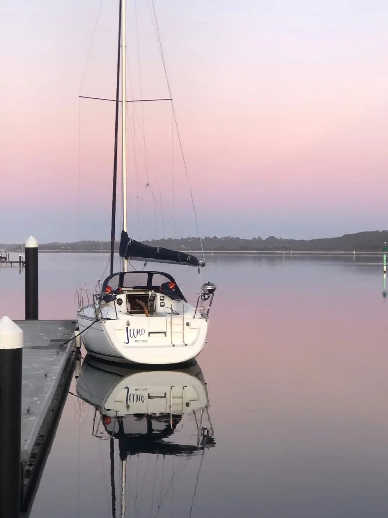 A sailboat named Jumo is docked at a calm marina during sunset, with pastel pink and purple sky reflected in the still water.