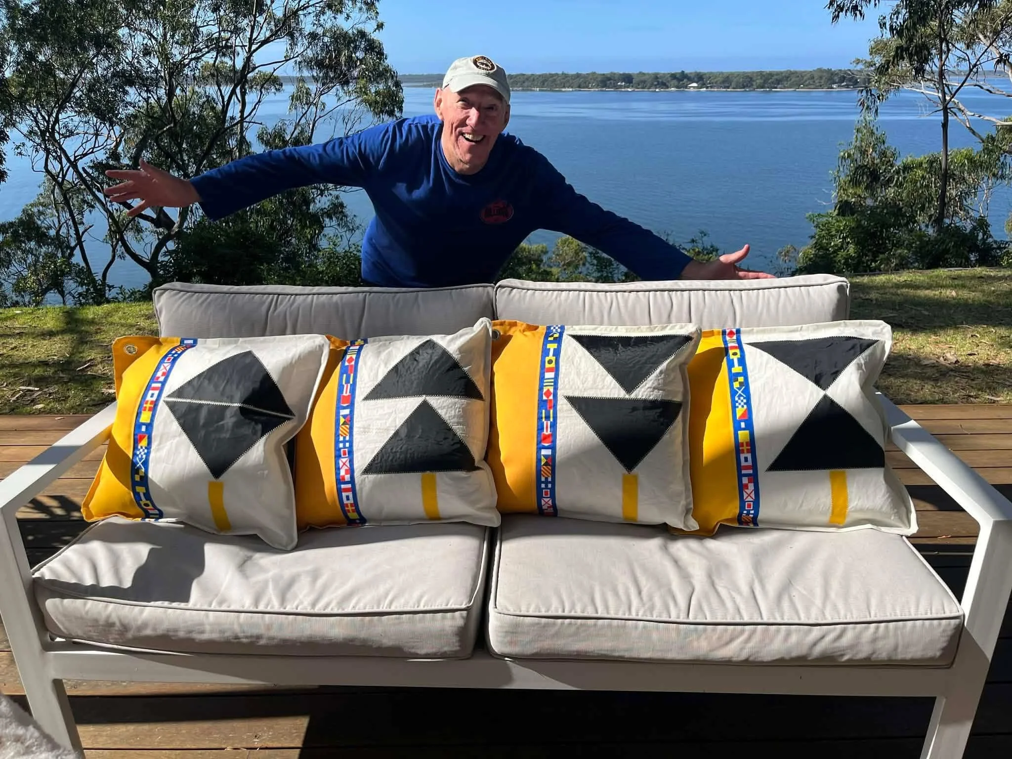 A man with a white cap and blue shirt is smiling and gesturing with his arms wide open behind a white outdoor couch with four decorative pillows featuring black and white geometric designs and a yellow background, overlooking a large body of water and trees on the horizon.