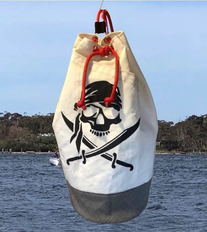 A white drawstring bag with a black skull and crossbones design, hanging above water with a boat and trees in the background.