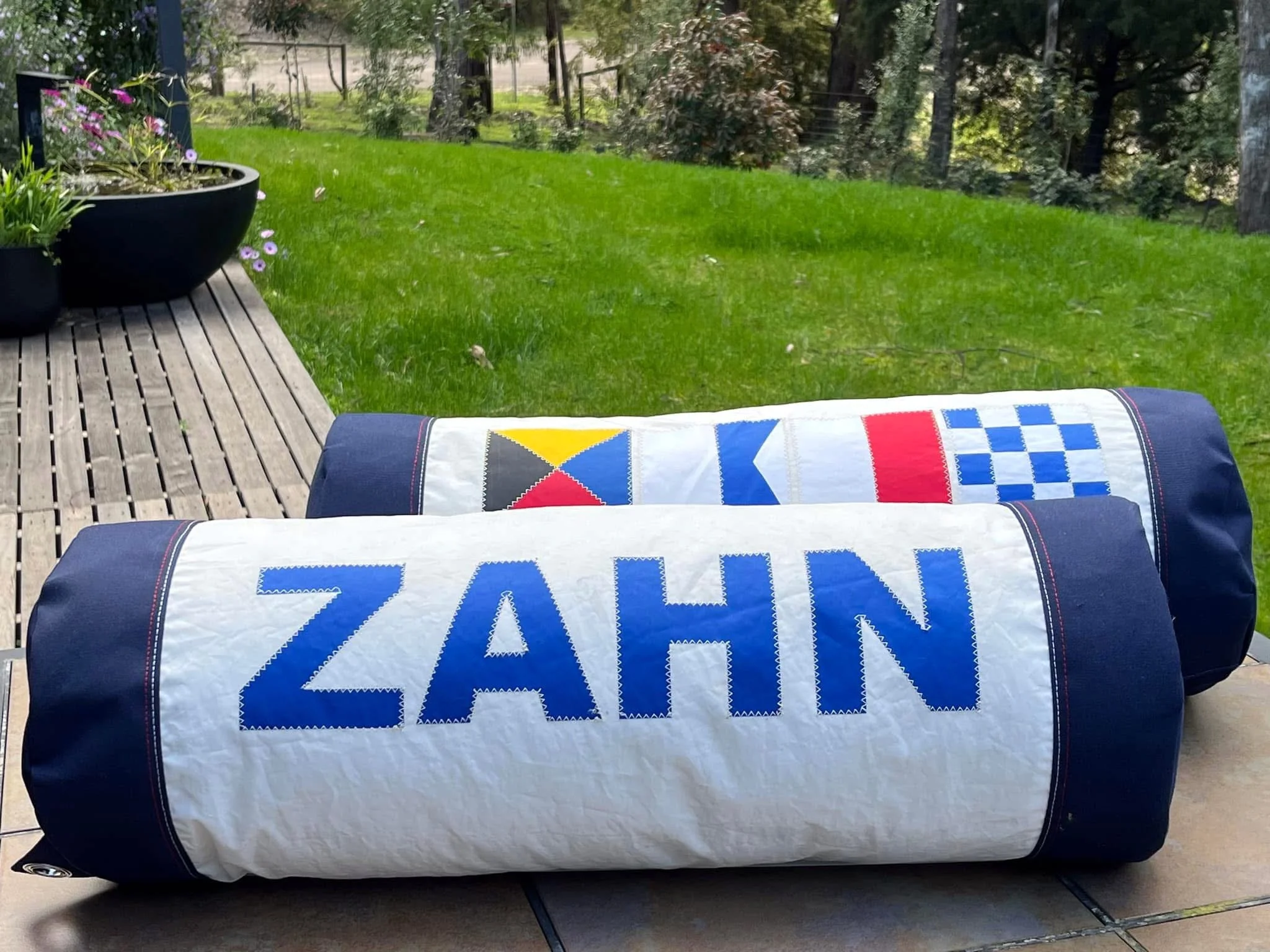Two cylindrical fabric-covered items with the name ZAHN printed on them, placed on a surface outdoors, with a grassy yard and trees in the background, and a potted plant with purple flowers nearby.