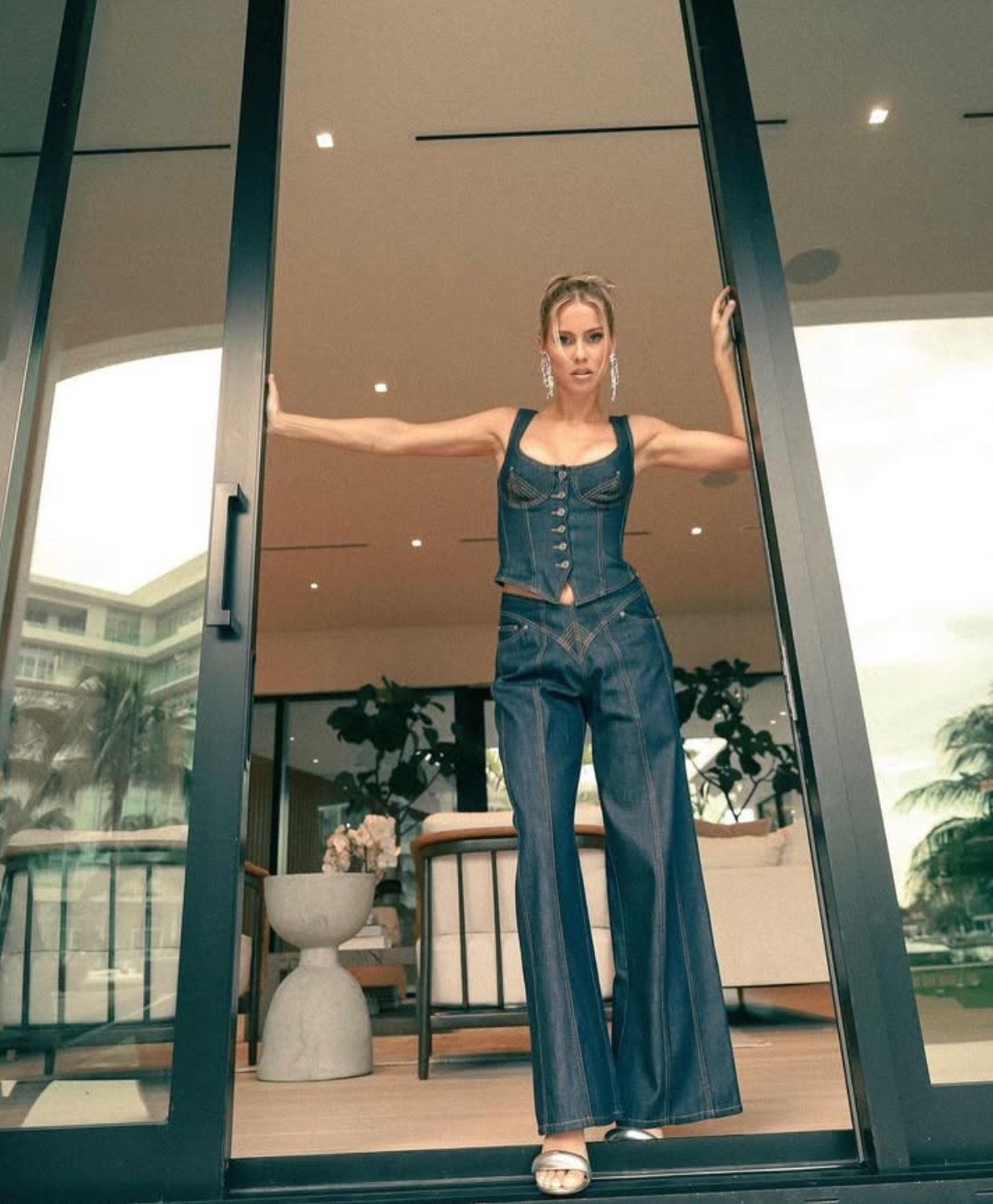 Person posing in a doorway wearing a denim corset top and wide-leg jeans, with outdoor furniture and plants visible in the background.