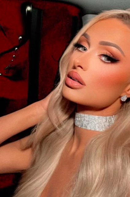 Portrait of a woman with long blonde hair, wearing a sparkling choker and earrings, posing in front of a red background.