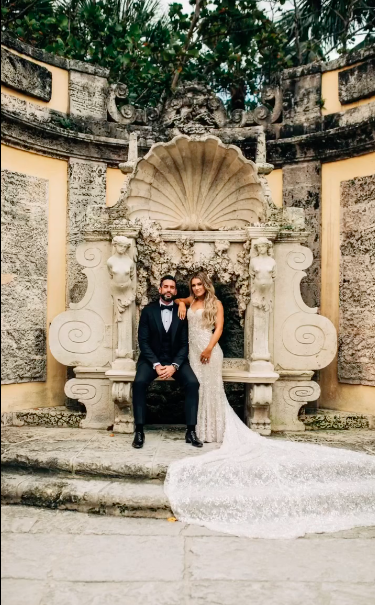 A couple in formal attire, a man in a suit and a woman in a wedding dress, posed in front of an ornate stone backdrop with a shell motif and cherub statues.