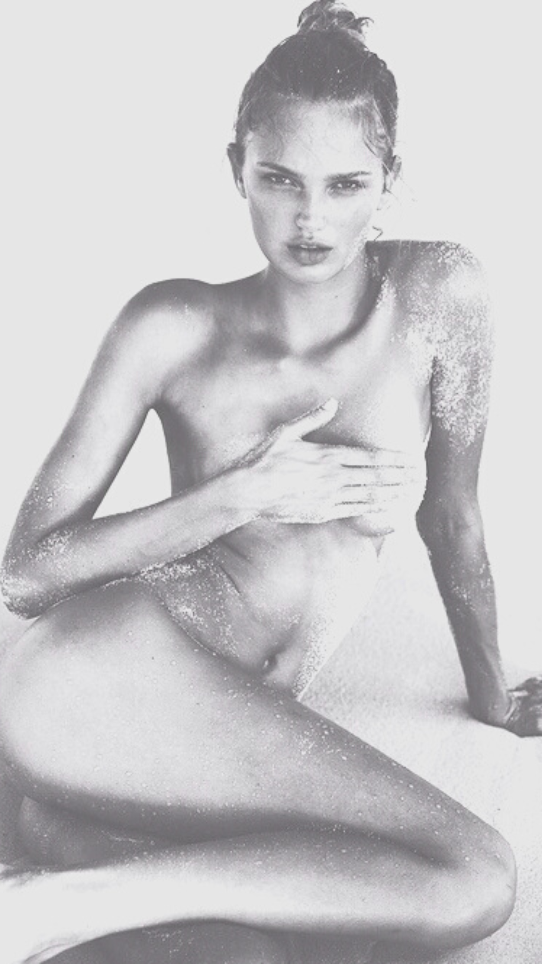 Black and white photograph of a seated person covered in sand, with one arm across the chest, and hair in a bun.