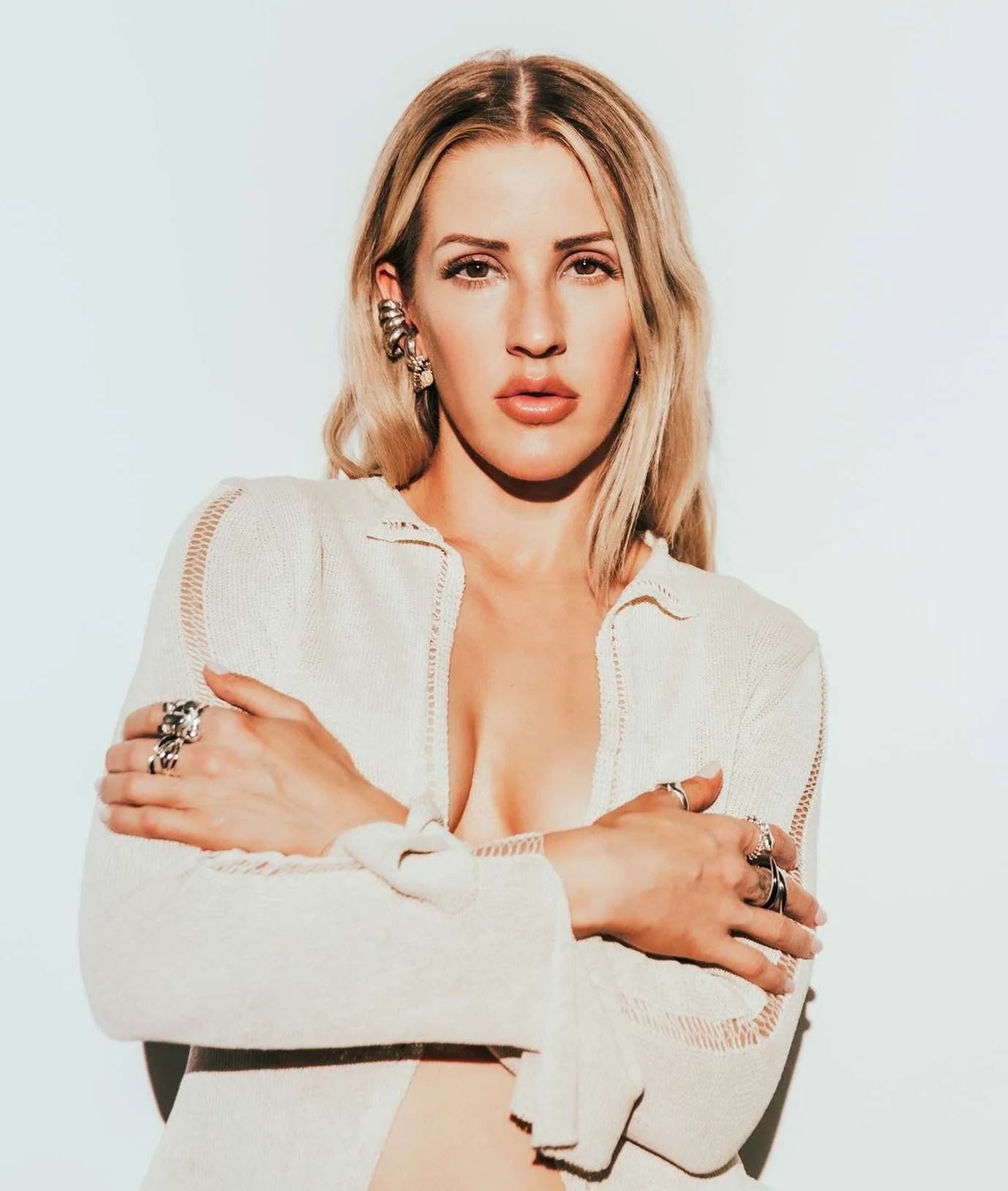 A woman with blonde hair wearing earrings and rings, crossing her arms, in a white cardigan against a plain light background. Ellie Goulding