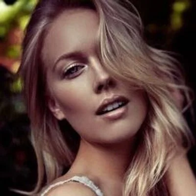 A woman with blonde hair and fair skin, partially covering one eye, posing outdoors with a blurred natural background.