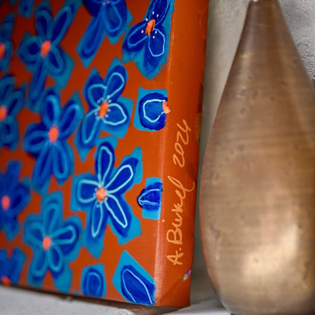 Close-up of a colorful floral painted canvas with the name 'A. Birkel 2026' written on the side, placed next to a smooth, curved clay vase.