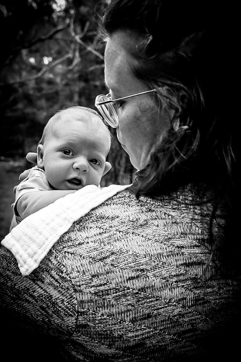 A woman holding a baby close to her shoulder outdoors, with the baby looking at the camera, in black and white.