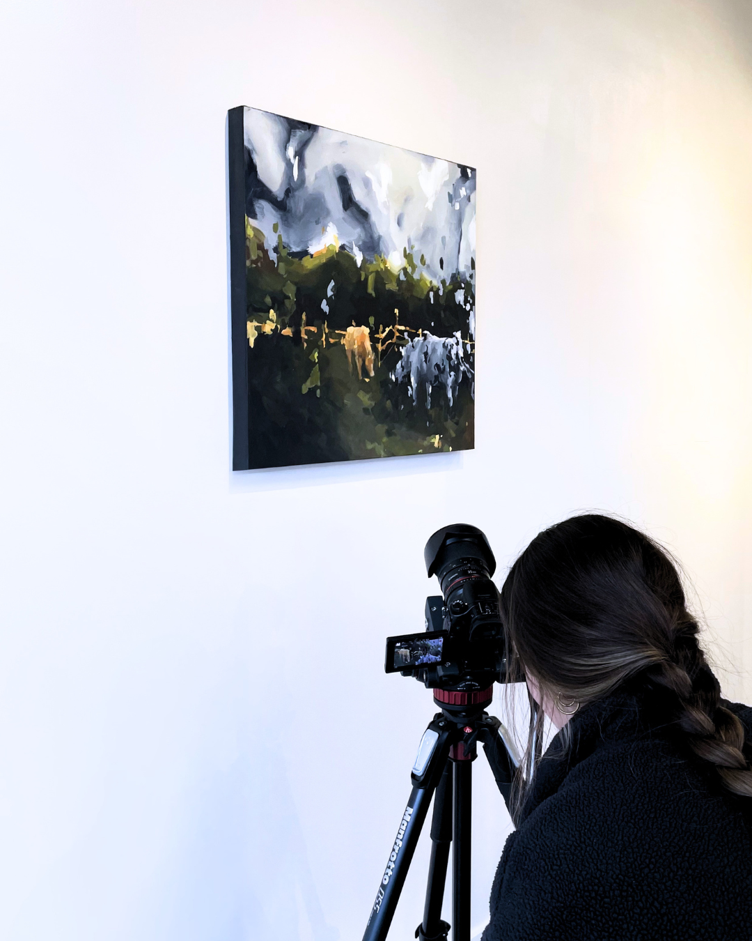 A woman with braided hair is taking a photo of a landscape painting hung on a white gallery wall using a professional camera on a tripod.