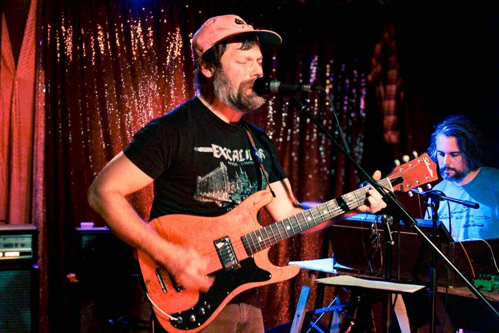 Colour photo of Smokey of Edmonton band Field + Stream playing guitar in front of the shimmery curtain on The Aviary stage. Guitar is red, and Smokey is wearing a pink hat and black t-shirt.