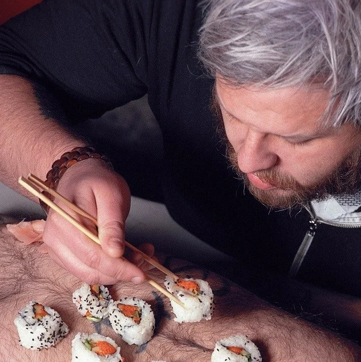 Craig Martell eats sushi off of a hairy belly with chopsticks. Colour photo, close up.
