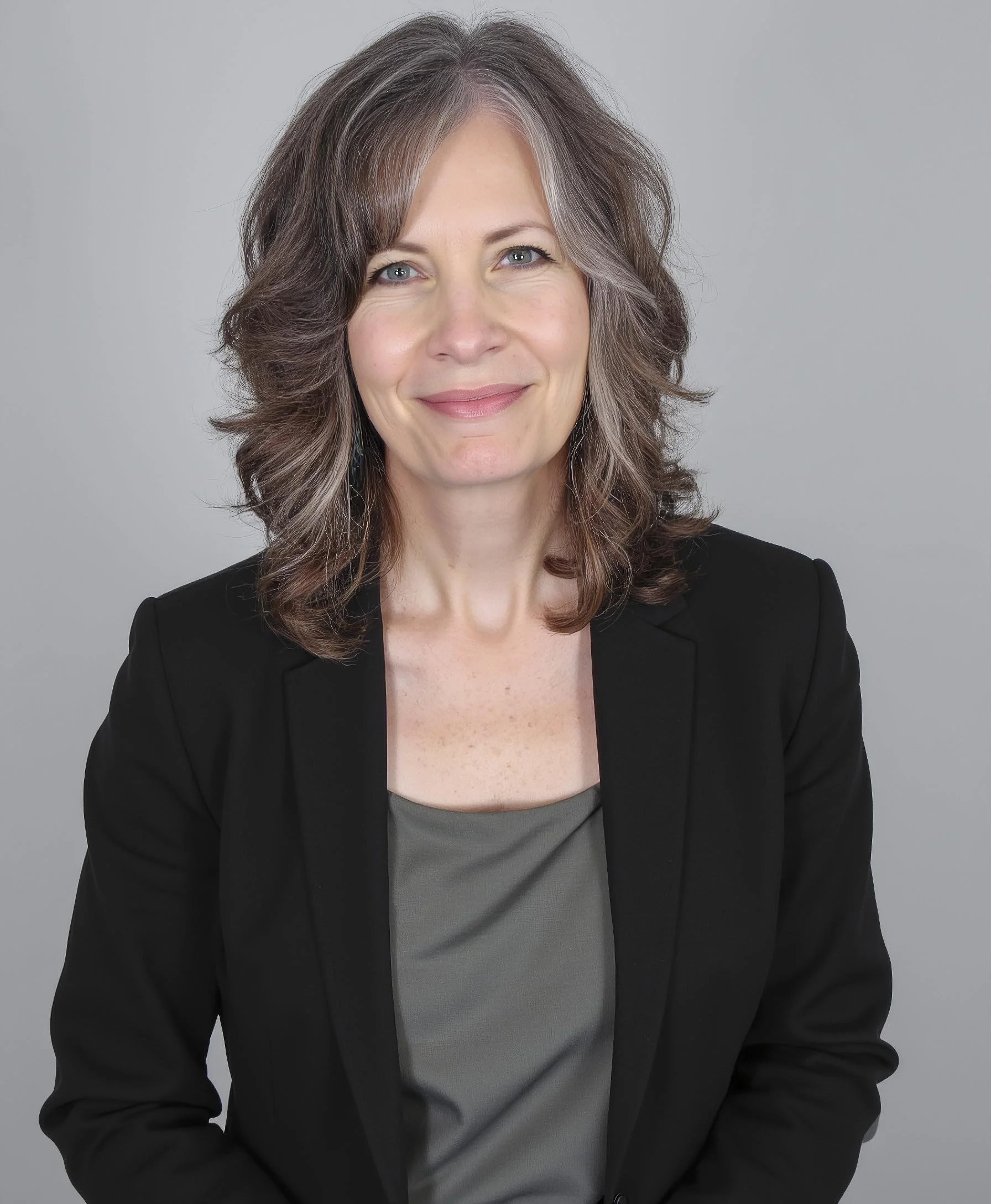 A woman with shoulder-length, wavy brown hair with gray streaks, wearing a black blazer and a gray top, smiling against a plain gray background.