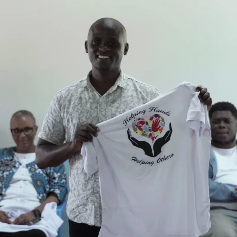 One of the community members smiling and holding a white T-shirt with a logo that says "Helping Hands Helping Others" at the inauguration of the Ndithini Vocational school.