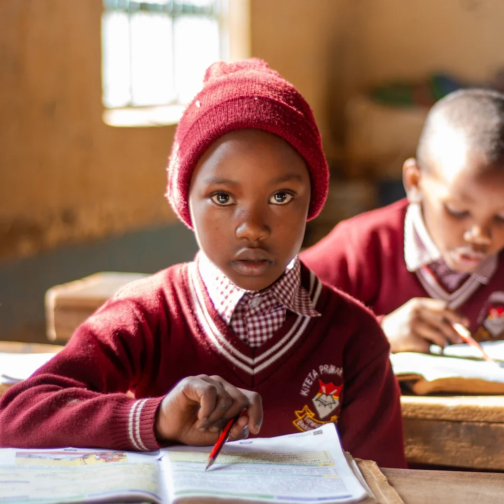 A Kiteta primary student wearing a red knit hat and school uniform, sitting at a wooden desk in a classroom and studying.