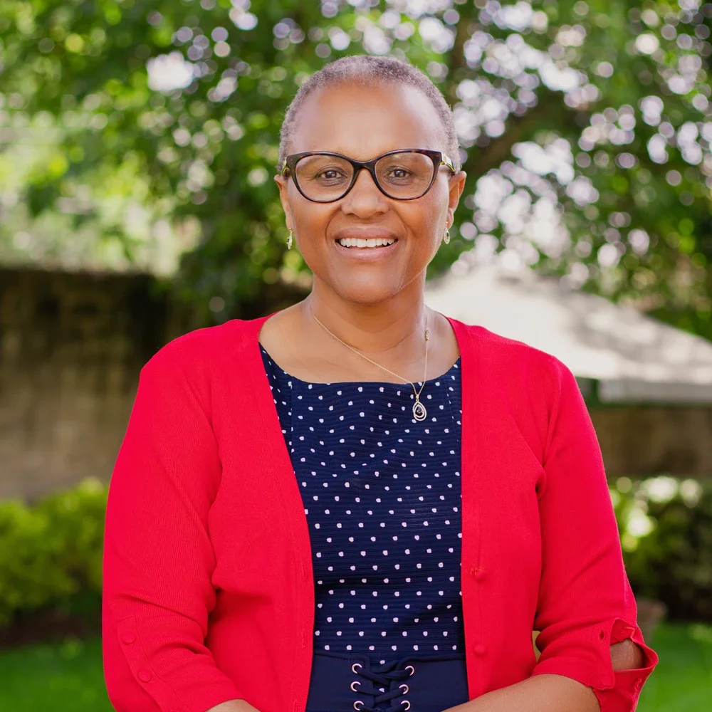 Pauline Mbuvi, Founder and President of Stephen Mbuvi Global, wearing glasses, a navy blue polka-dot shirt, a red cardigan, and jewelry, standing outdoors with blurred greenery in the background.