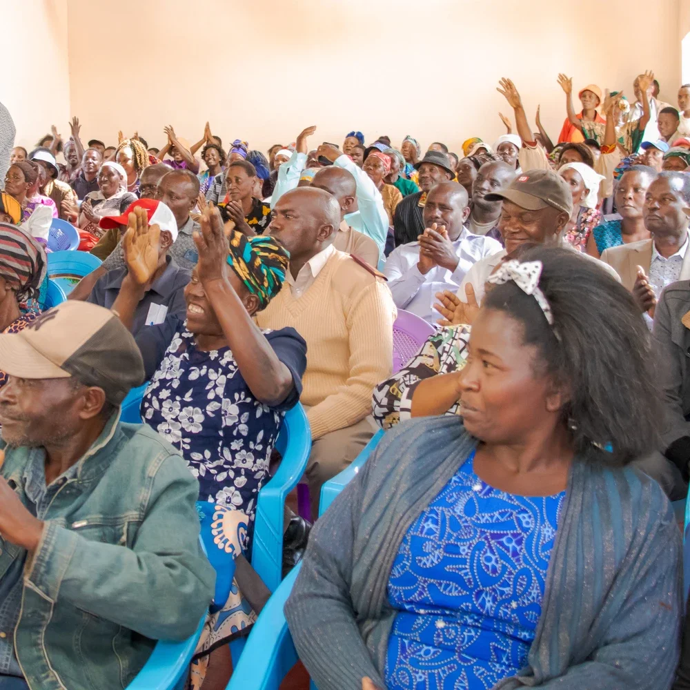 A large group of diverse people at an indoor event, some clapping and raising their hands in celebration, with smiles and attentive expressions.