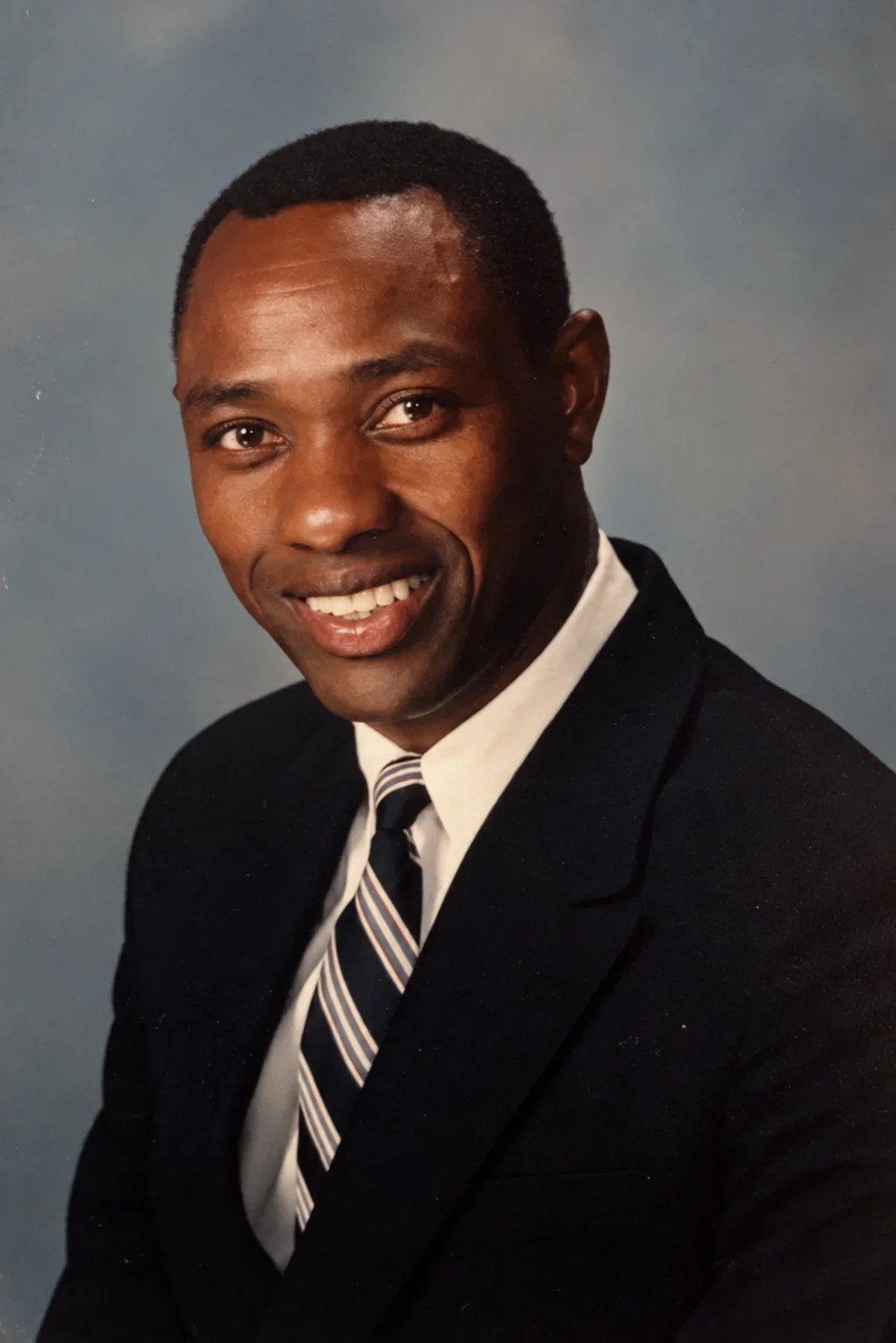 Dr. Stephen Mbuvi, whose legacy inspired Stephen Mbuvi Global, in a professional portrait wearing a black suit, white shirt, and a striped tie, smiling and looking at the camera against a plain gray background.