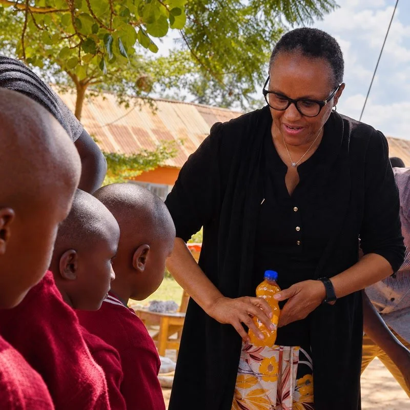 Pauline Mbuvi sharing a meal with children during a community visit in Kiteta Primary School, interacting with children, holding a yellow bottled drink in the outdoors.