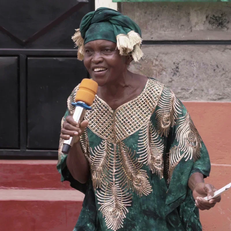 A local community leader wearing a green headscarf and traditional African attire holding a microphone and addressing the gathering during the inauguration of Ndithini Vocational school.