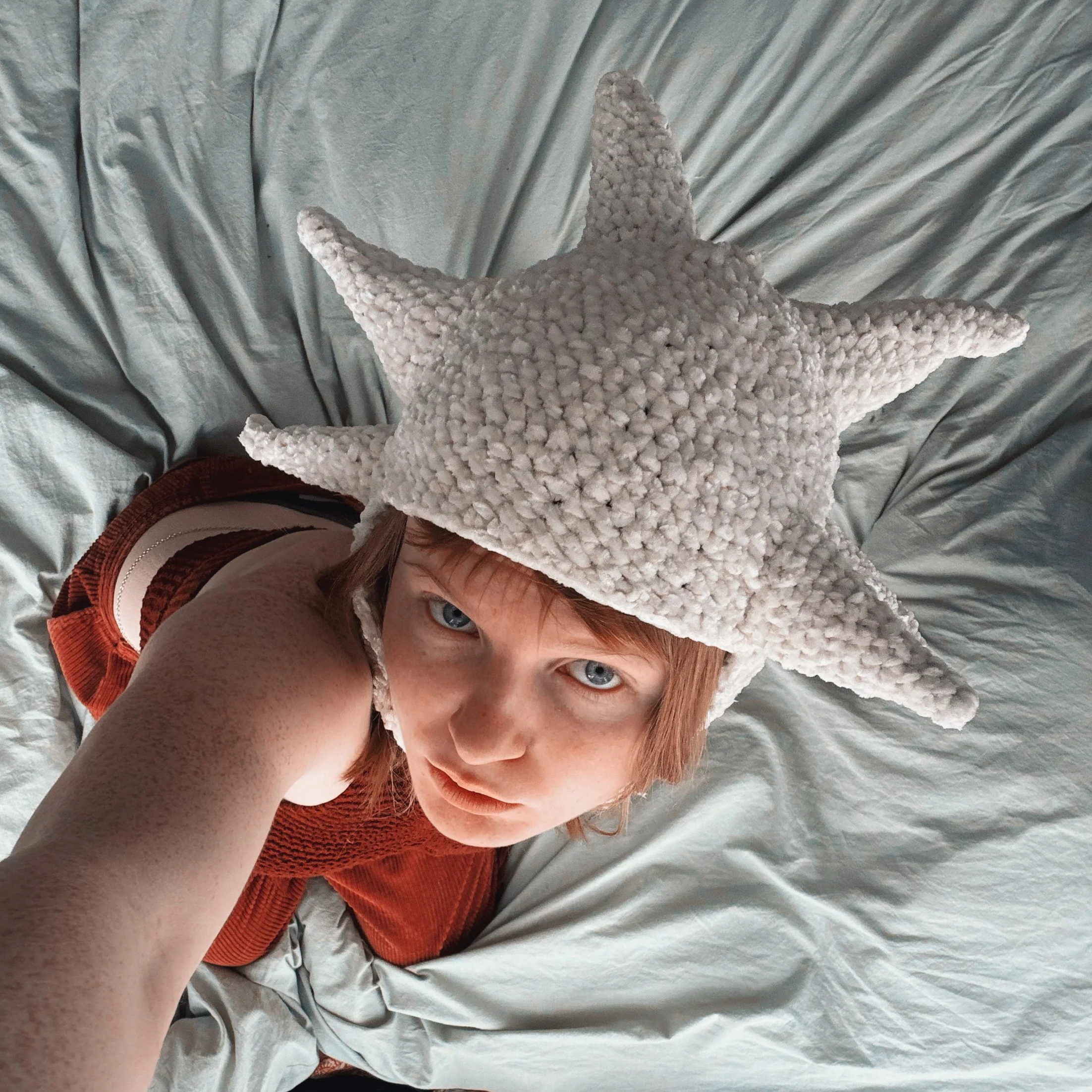 A person with blue eyes and red hair wears a large white starfish-shaped hat, lying on a wrinkled gray sheet.