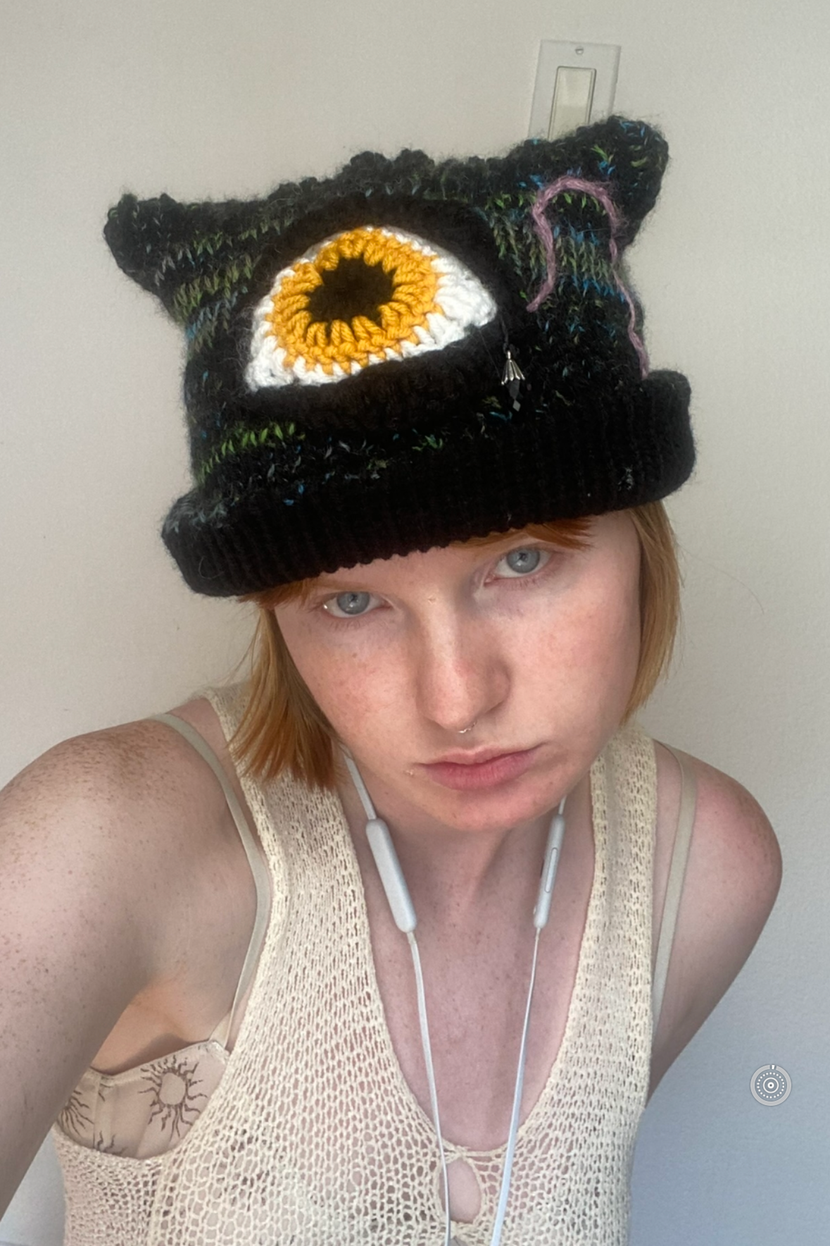 Person with red hair and freckles wearing a crochet hat with a large eye design, a beige mesh top, and white earphones.
