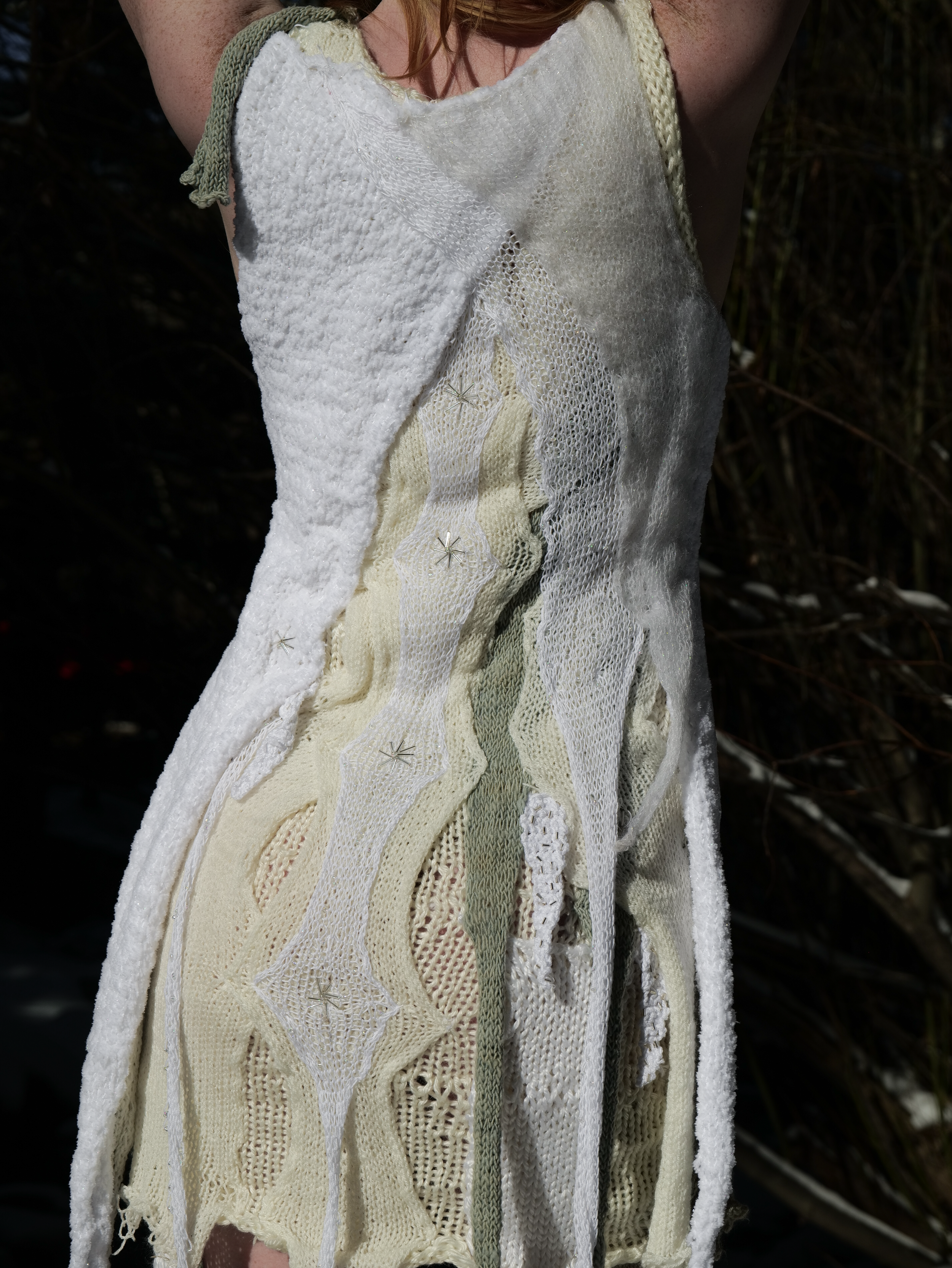 Close-up of a dress made from various patterned and textured fabric patches in cream, white, and pale green, pinned together with small metal pins.