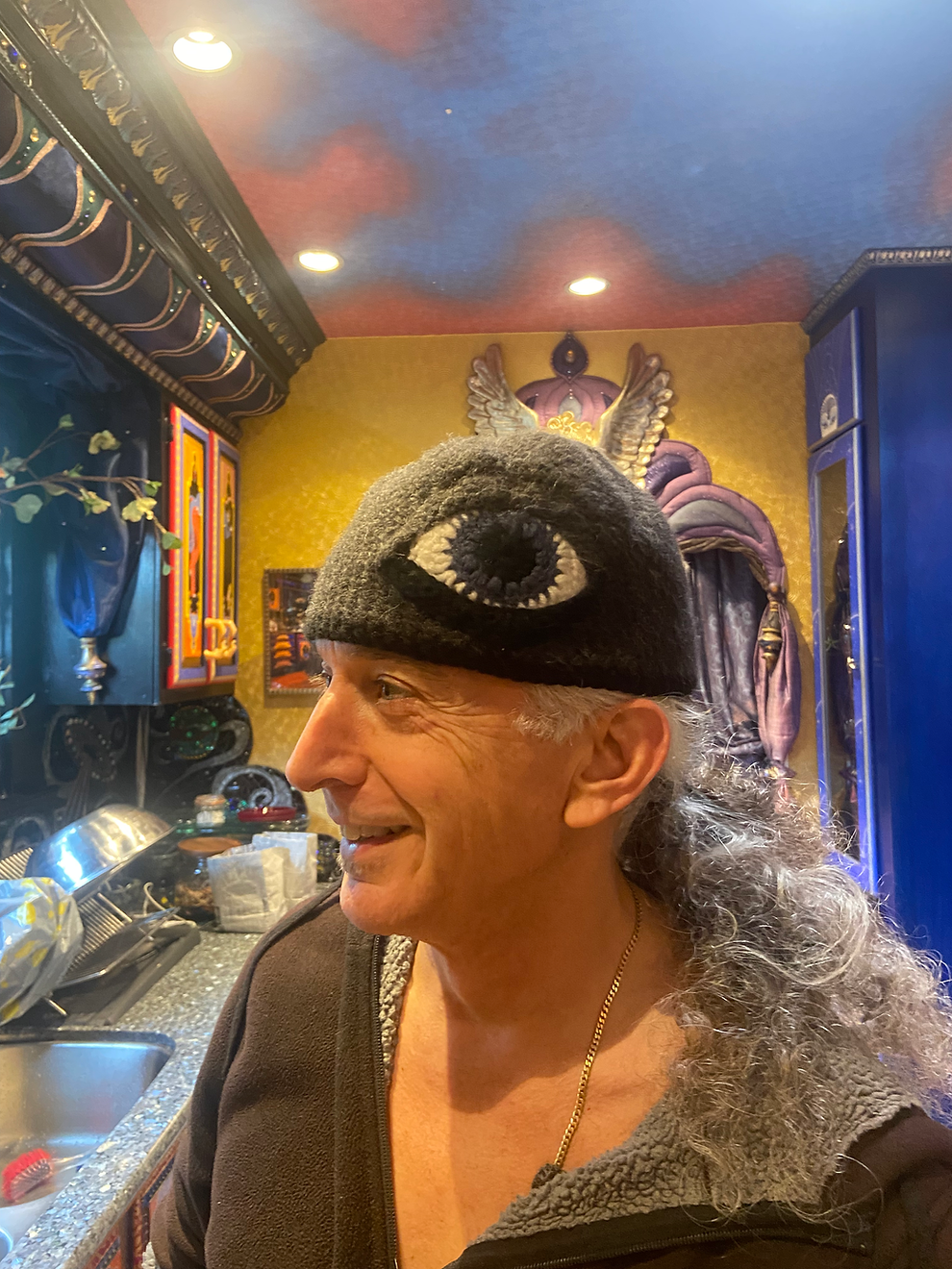 A man with long gray curly hair wearing a black hat with a white eye design, smiling in a colorful, decorated room.
