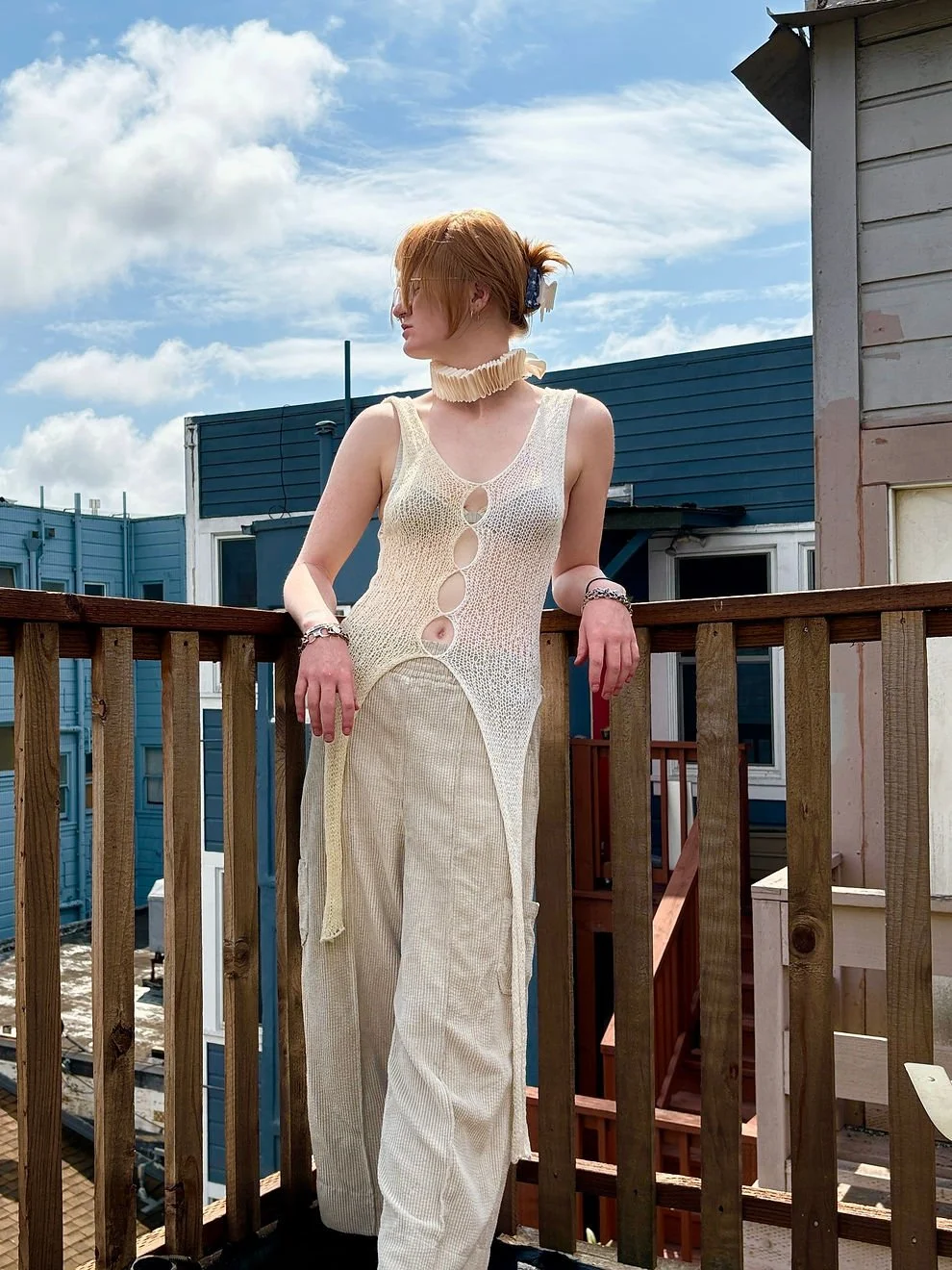 A young woman with short red hair wearing a cream-colored, sleeveless, fishnet dress with large circular cutouts, and wide-leg trousers, standing on a balcony with wooden railing against a blue sky with clouds.