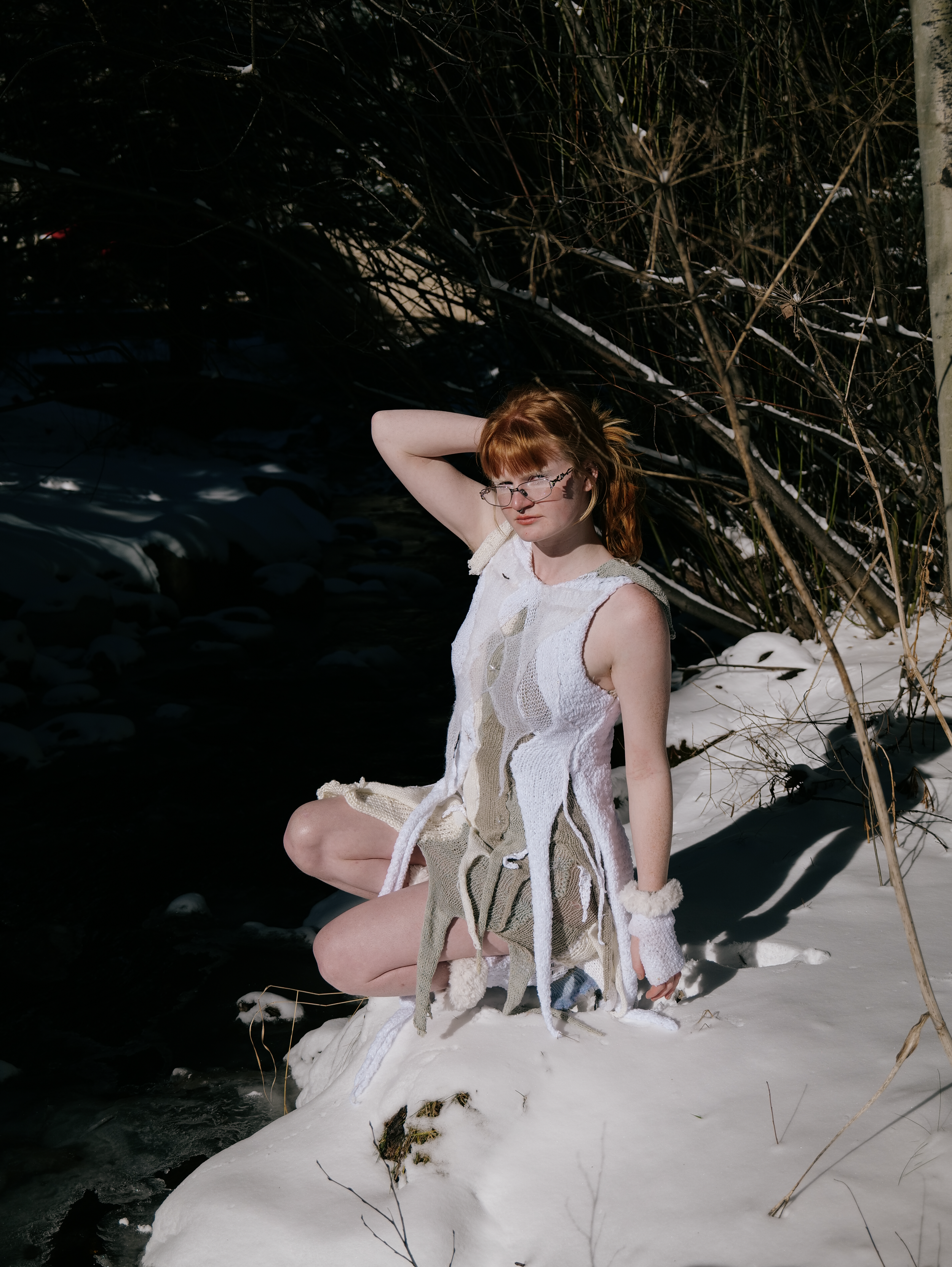 A young woman with red hair and glasses kneels in the snow beside a creek, wearing a sleeveless, handmade, multicolored knitted dress, with bare legs and a serious expression, surrounded by leafless trees and bushes.