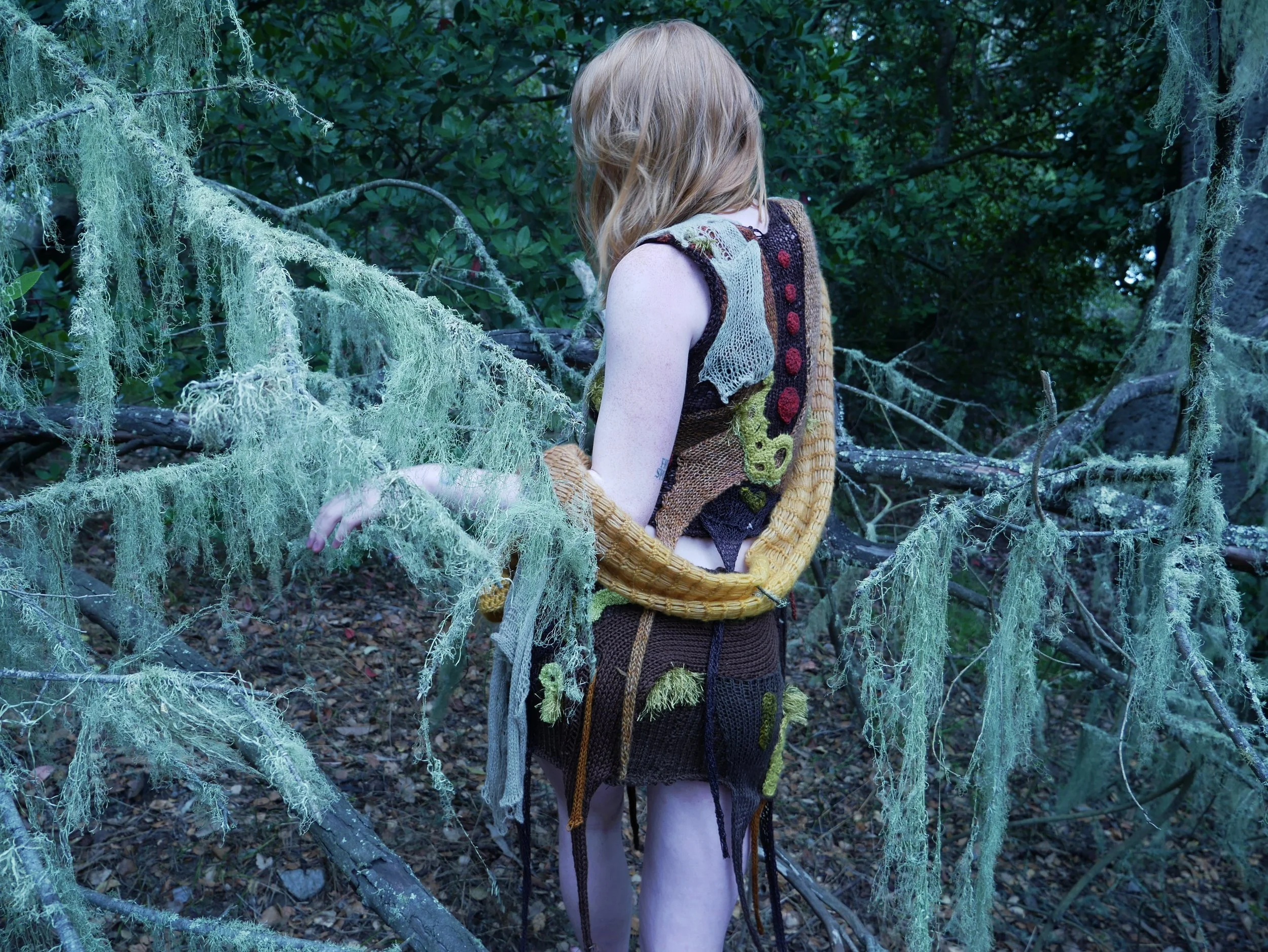 A young woman with light brown hair standing in a wooded area, surrounded by tree branches covered in moss. She wears colorful knitted clothing and carries a woven basket on her back.