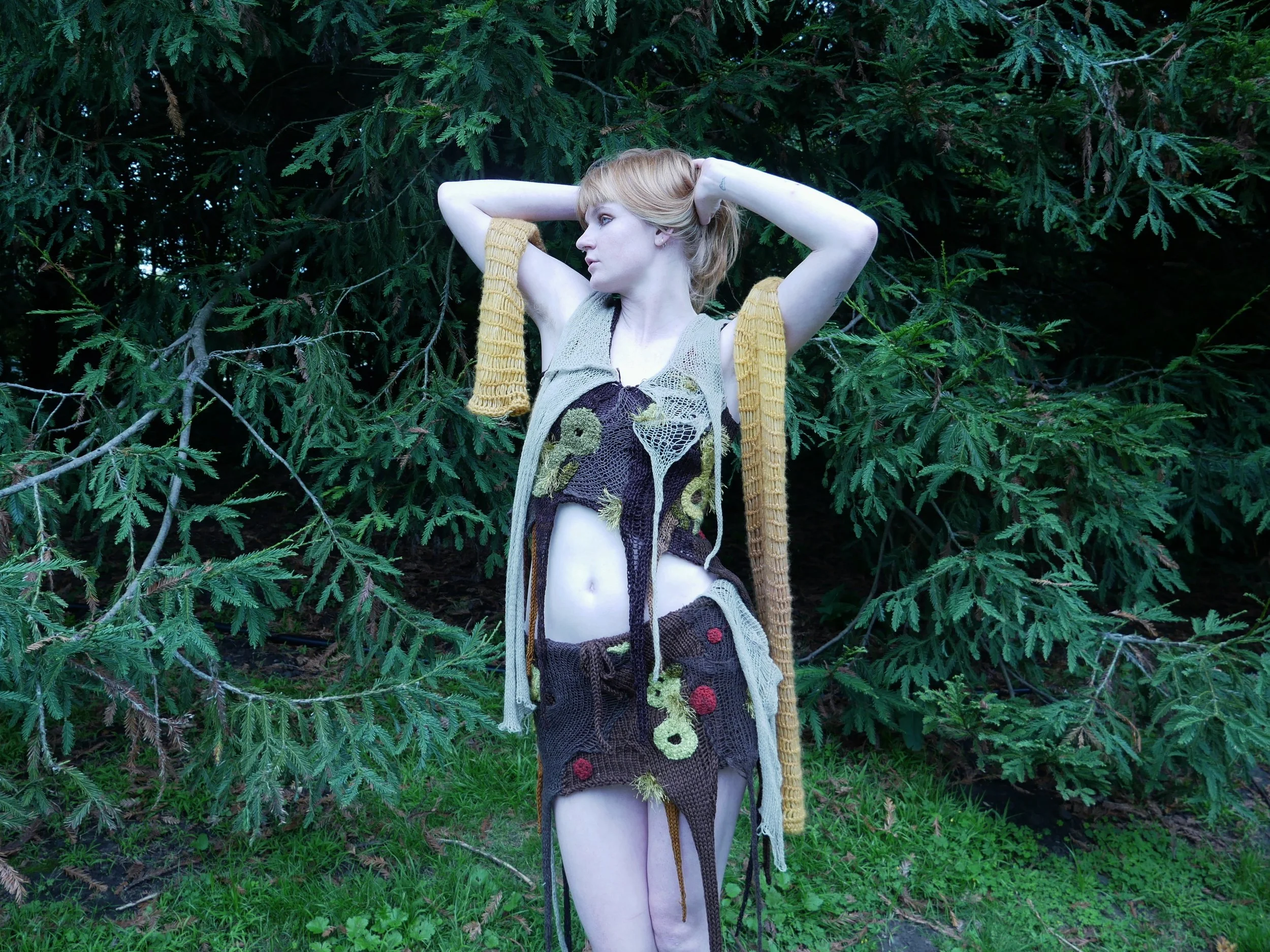 A young woman with short red hair standing outdoors in front of dense green foliage, wearing a handmade, eclectic outfit with knitted and crochet elements in earthy tones.