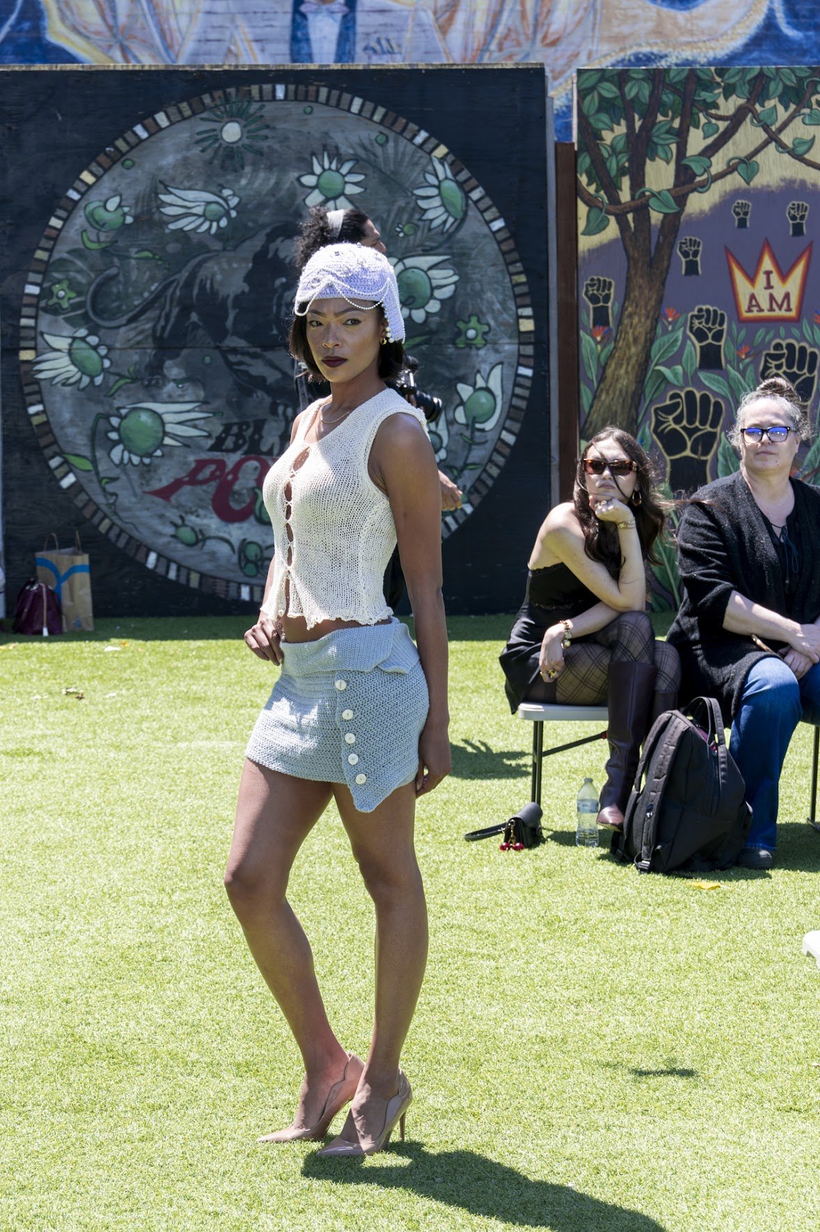 A woman in a white sleeveless crochet top and matching mini skirt with buttons, wearing high heels, posing outdoors on a grassy lawn during daytime. Behind her, two women are seated, one with sunglasses and the other with glasses, resting on a folding chair, with colorful murals and art in the background.