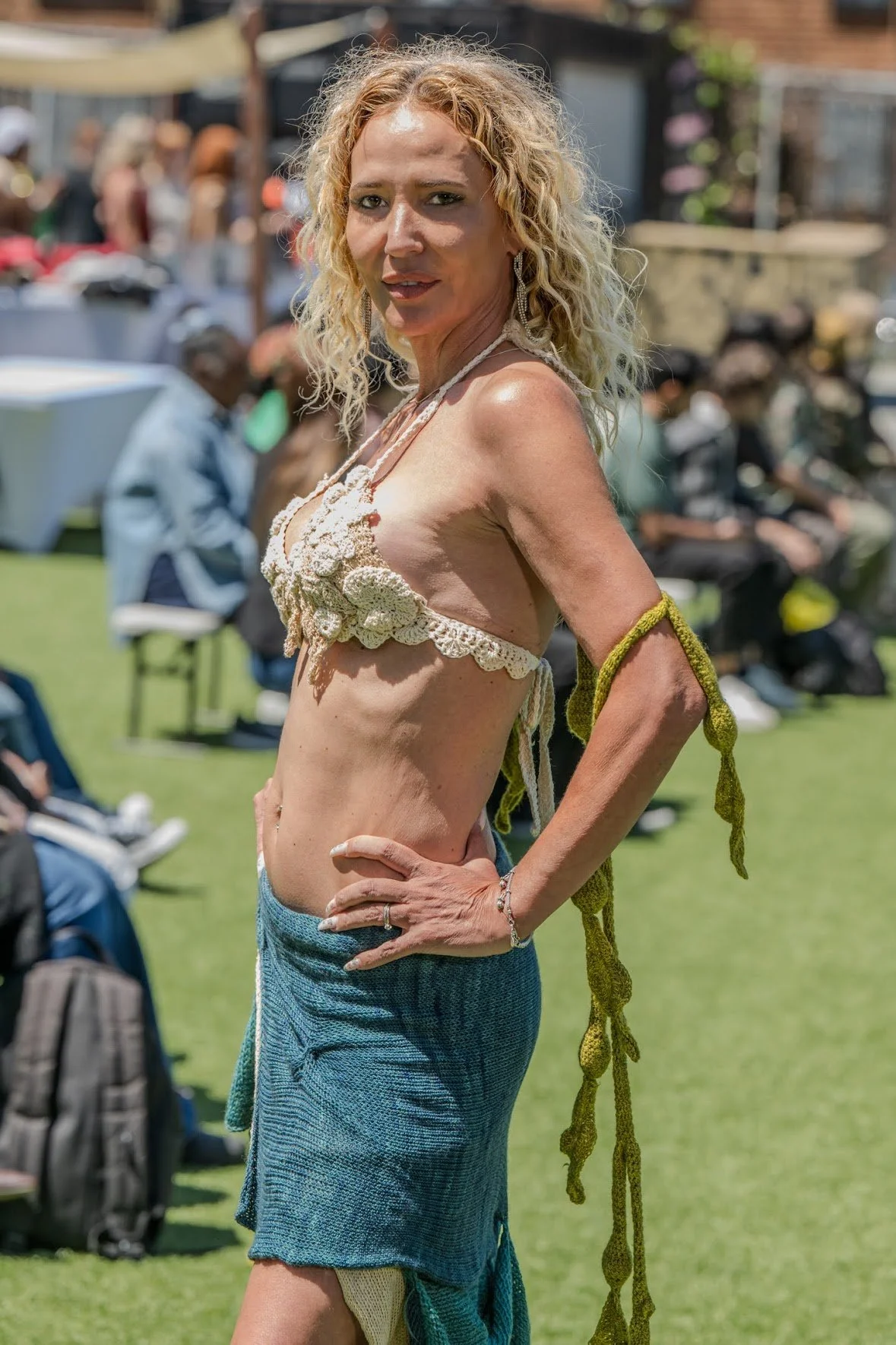 A woman with curly blonde hair wearing a crocheted top and a long skirt outdoors, with people and chairs in the background.