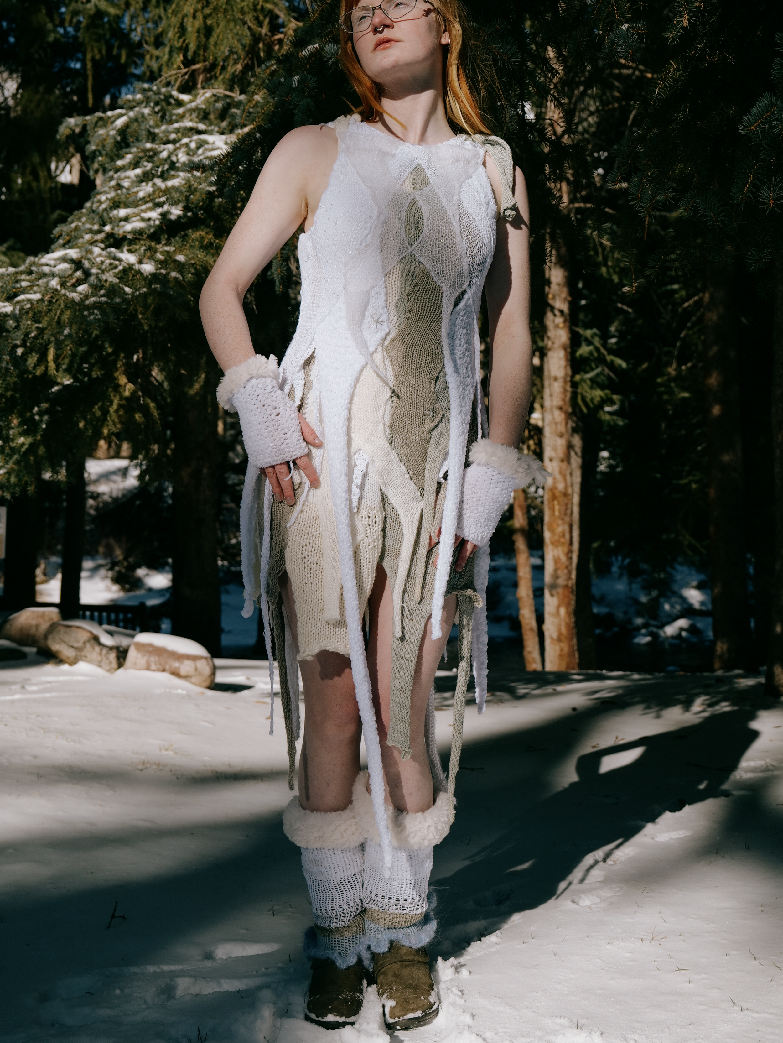 A woman with red hair dressed in a handmade, knitted dress with long, loose fabric strips, standing outdoors in the snow with evergreen trees in the background.
