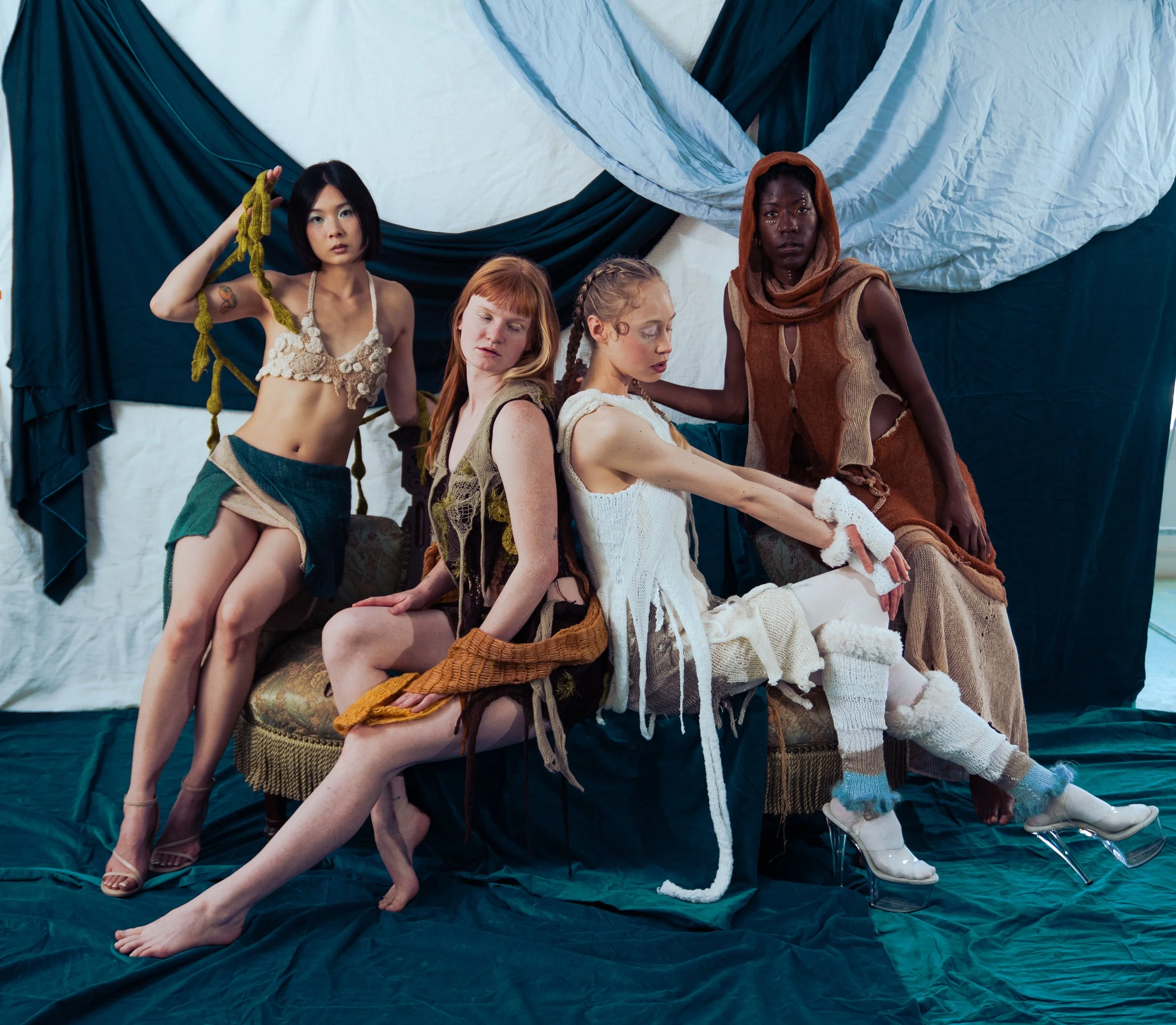 Group of five diverse women in eclectic clothing sitting and posing against a background of dark and light draped fabrics.