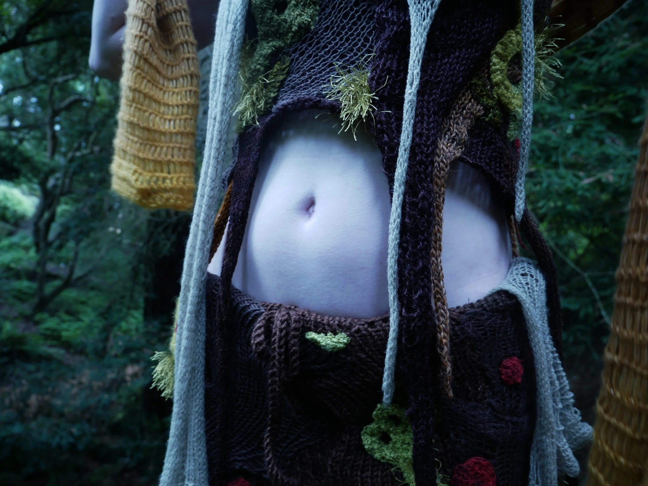 Close-up of a person's midsection with a crop top, surrounded by colorful knitted and crocheted fabrics with floral decorations, outdoors near trees.