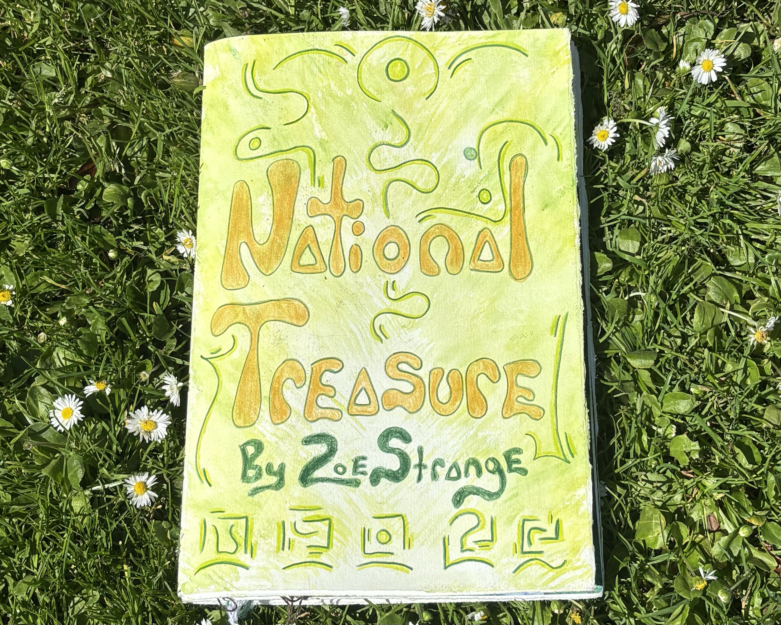 Handmade sign on green paper with yellow, orange, and green writing and drawings, placed on grass with small white daisies.