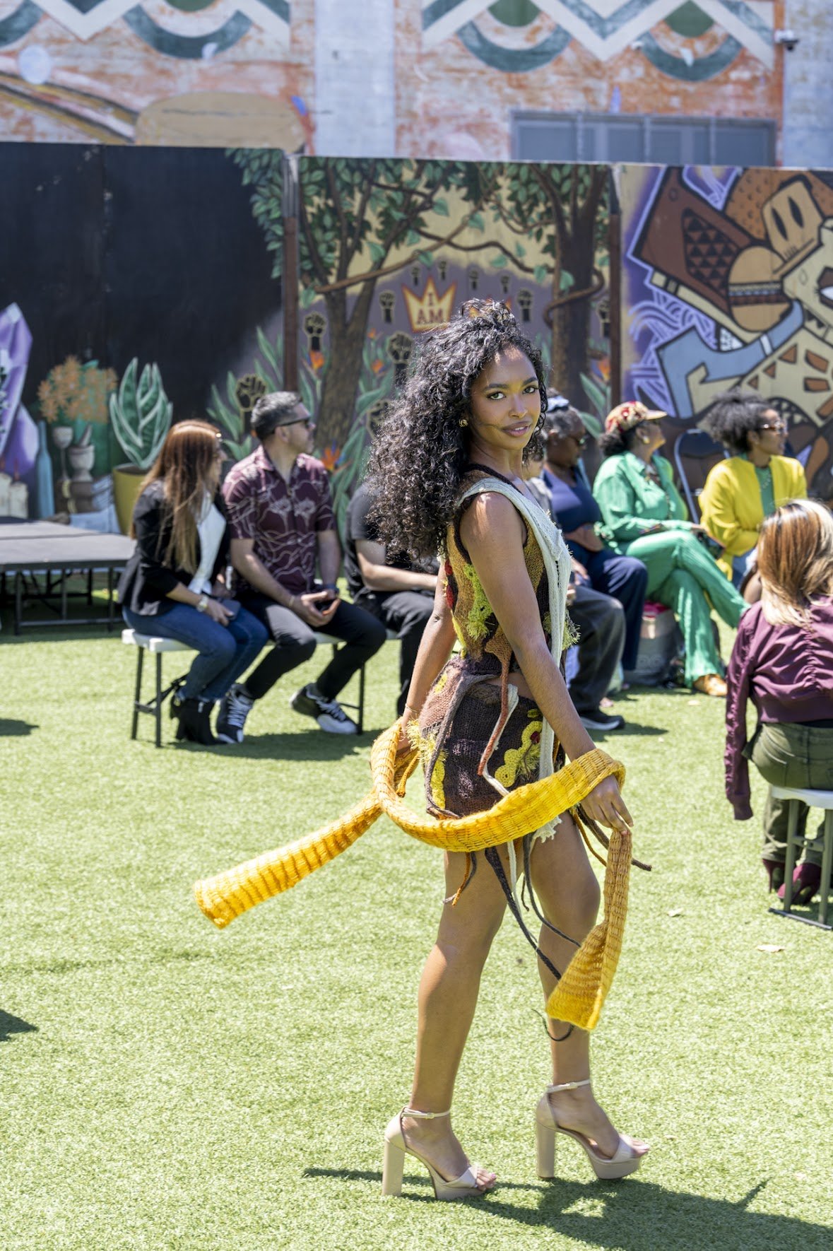 A woman with curly hair wearing a colorful, artistic outfit with beige high heels, walking on a grassy area at an outdoor event with a seated audience in the background.