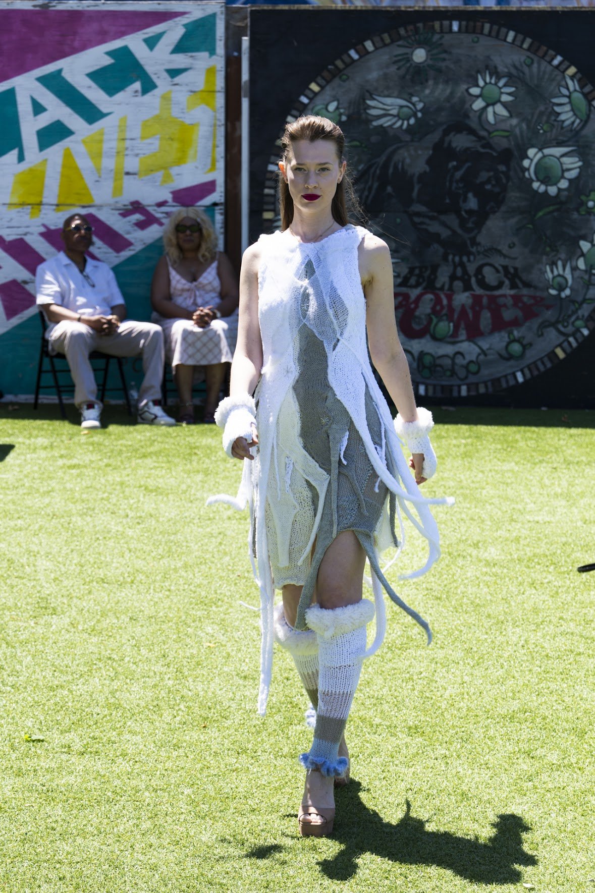 A woman walking on a grassy field during a fashion event, wearing a unique white and gray knitted dress with fur trim and platform heels, with two seated spectators and colorful murals in the background.