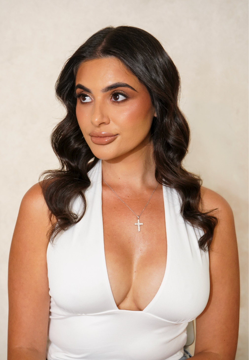 A woman with dark, wavy hair wearing a white sleeveless top and a silver cross necklace.