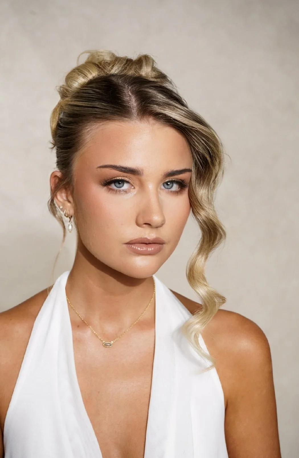 Close-up portrait of a young woman with blonde hair styled in loose waves, blue eyes, and natural makeup, wearing gold jewelry, a white halter top, and gazing at the camera against a neutral background.
