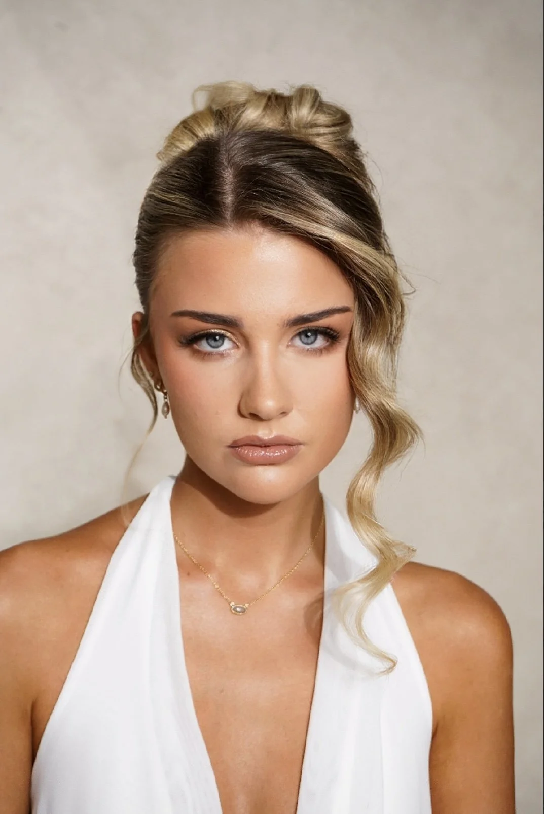 A young woman with blonde hair styled in a side-swept wave, wearing a white sleeveless top, gold necklace, and earrings, looking directly at the camera with neutral expression.