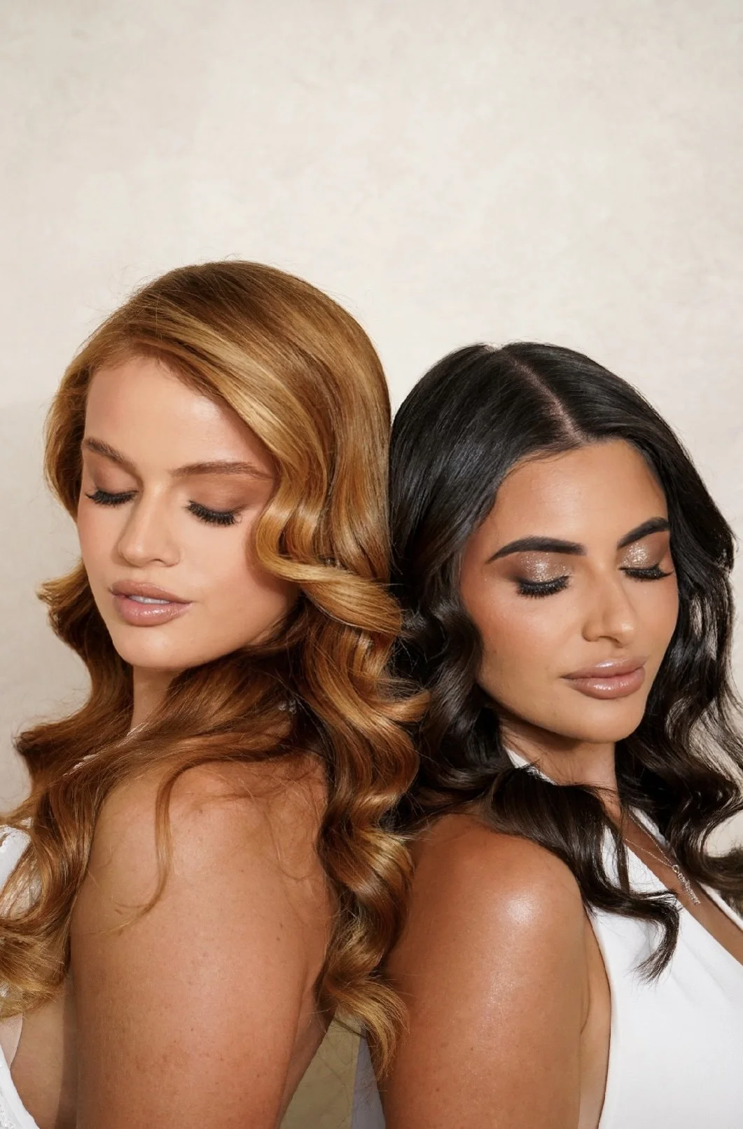 Two women with long, wavy hair and makeup, standing back to back with eyes closed against a plain background.