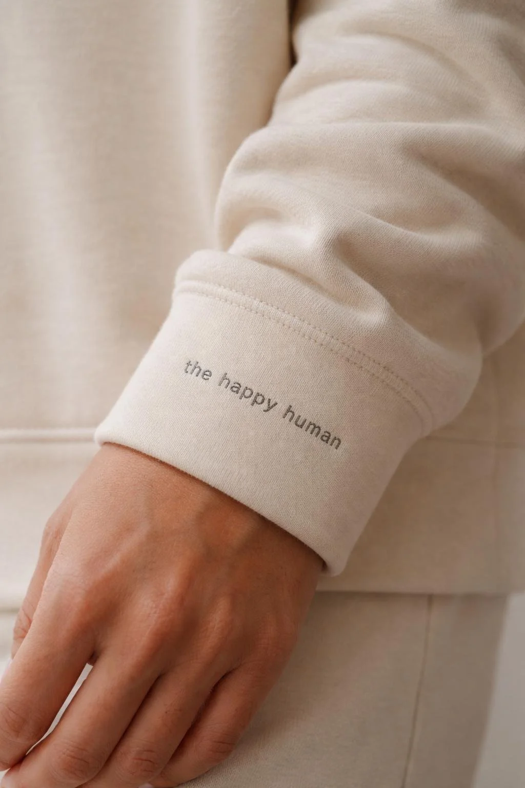 Close-up of a person wearing a beige sweatshirt with the words "the happy human" embroidered on the cuff of the sleeve.
