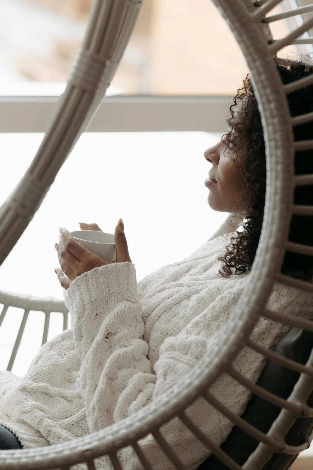 A woman with curly hair, wearing a white knit sweater, sitting in a rattan chair, holding a white mug, in a cozy indoor setting with natural light.