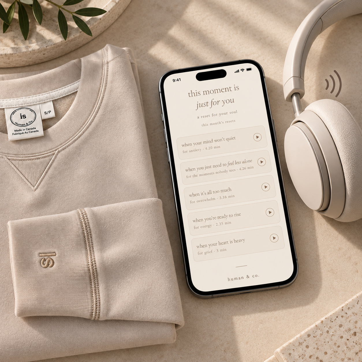 A flat lay of a beige sweatshirt, a smartphone with a meditation app open, and a pair of white over-ear headphones on a beige surface.
